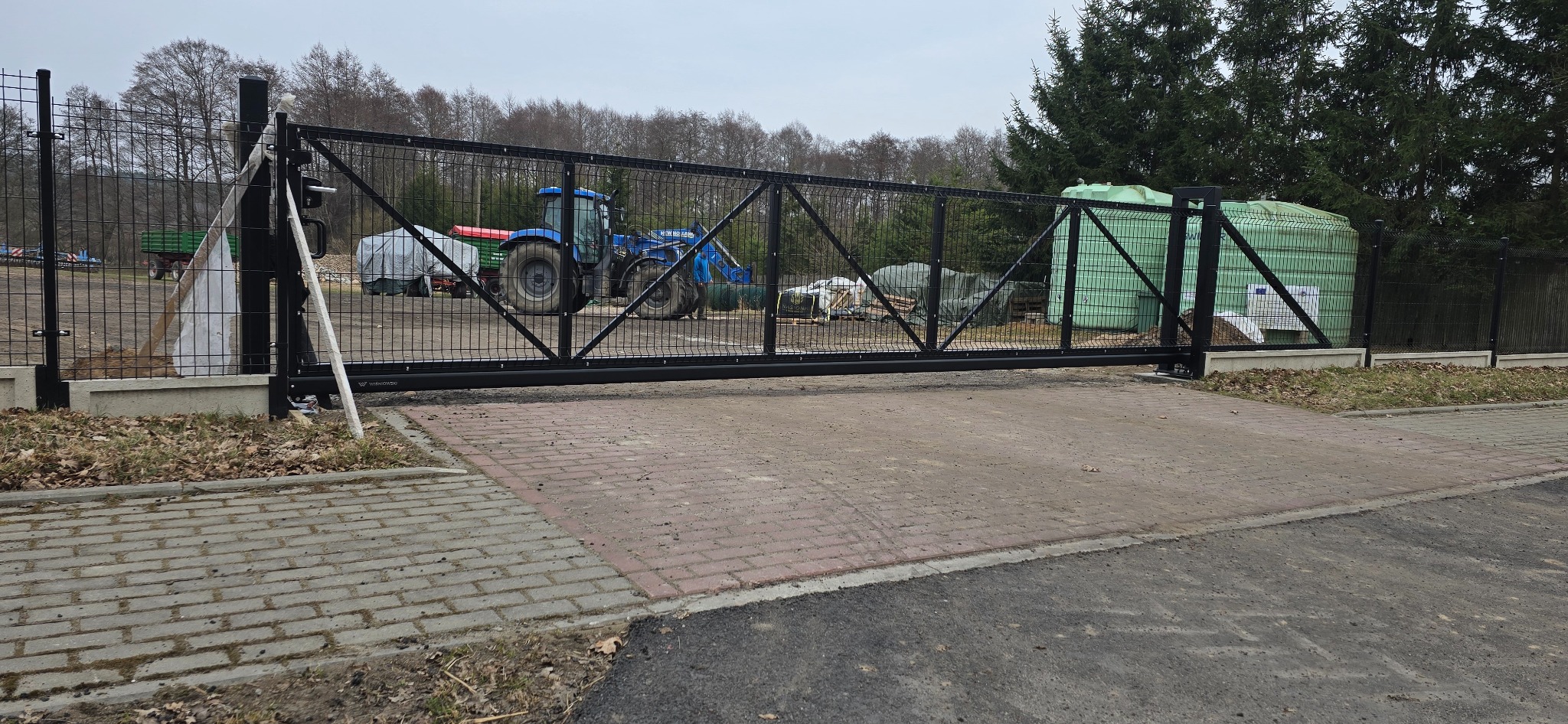 Czarna, przesuwna brama wjazdowa z metalowej siatki na posesji. W tle widoczny traktor i zbiorniki na wodę. Brama zamontowana na betonowym fundamencie.