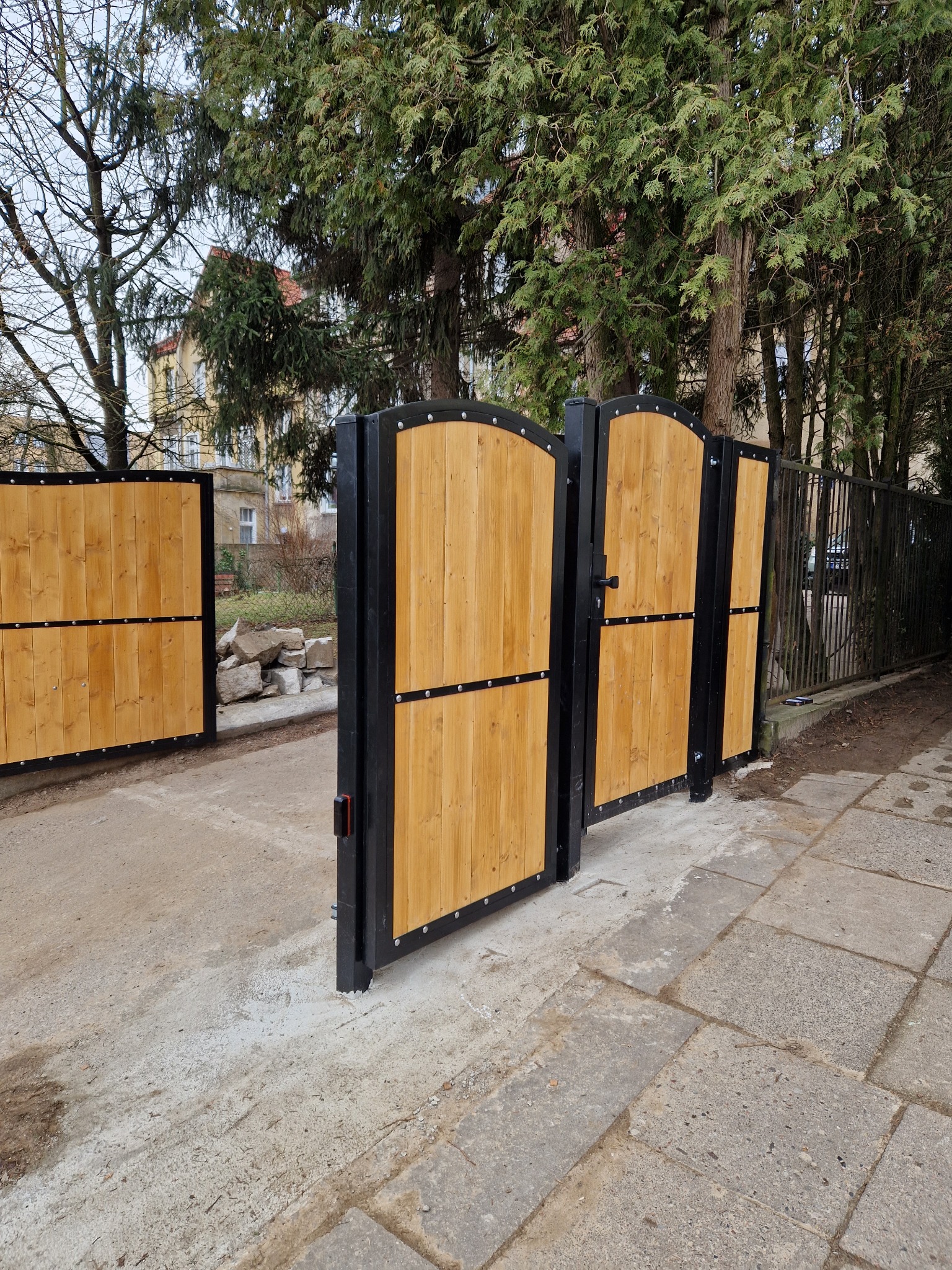 Drewniana brama wjazdowa z czarną metalową ramą, zamontowana w Kołbaskowie, widoczna z boku, z fragmentem ogrodzenia panelowego w tle.