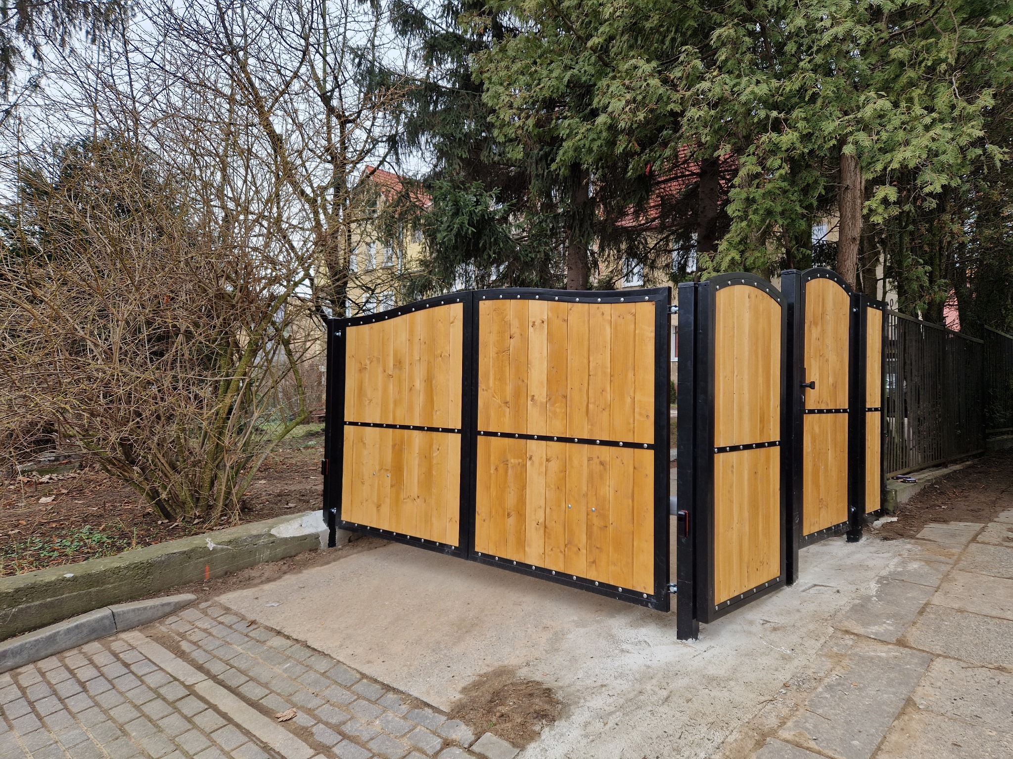 Dwuskrzydłowa brama wjazdowa z jasnego drewna i czarną metalową ramą, widoczna z zewnątrz posesji, na tle drzew i częściowo widocznego budynku.