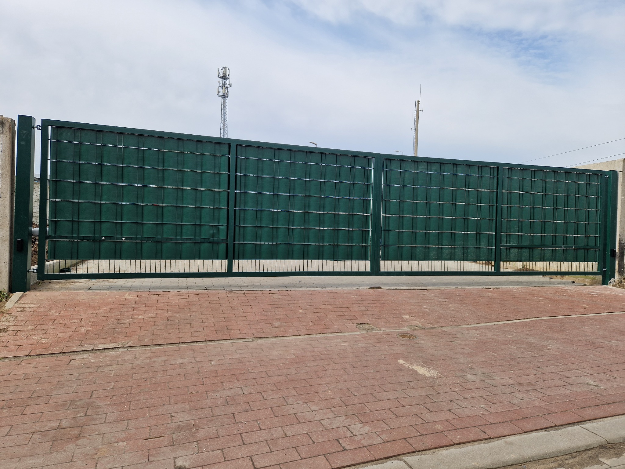 Zamykana, przesuwna brama wjazdowa z zielonych paneli z drobną siatką, na posesji wyłożonej czerwoną kostką brukową, w tle maszt telekomunikacyjny.