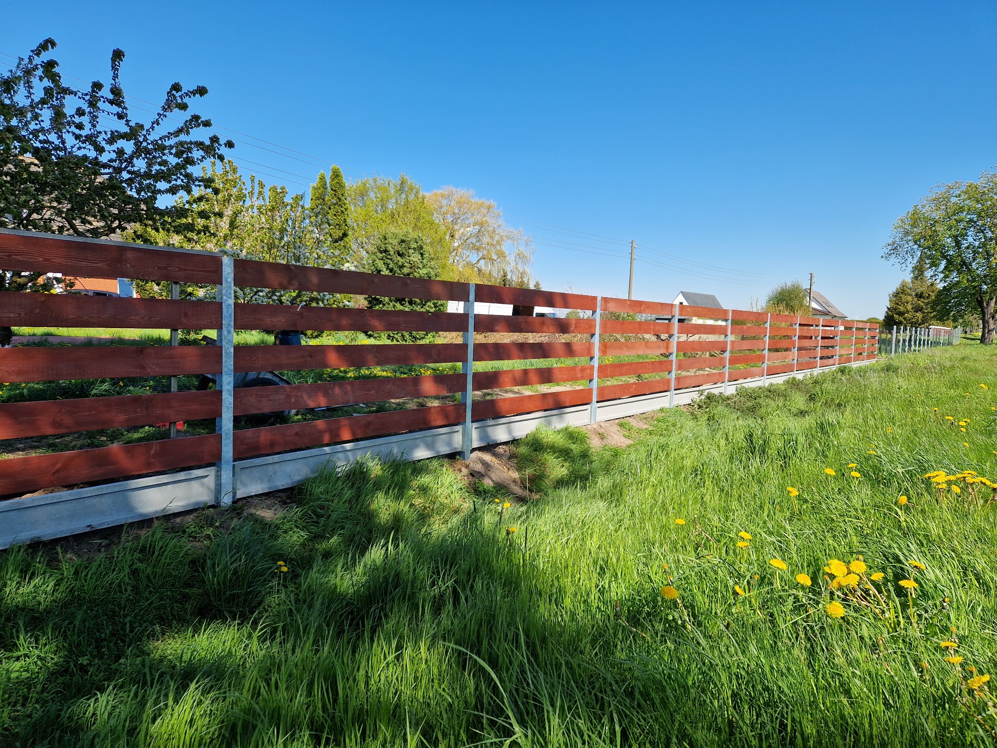 Długie, horyzontalne ogrodzenie z brązowych, drewnianych desek, wsparte na metalowych słupkach i betonowej podmurówce, ciągnące się wzdłuż zielonej łąki z kwitnącymi mniszkami, pod błękitnym niebem.