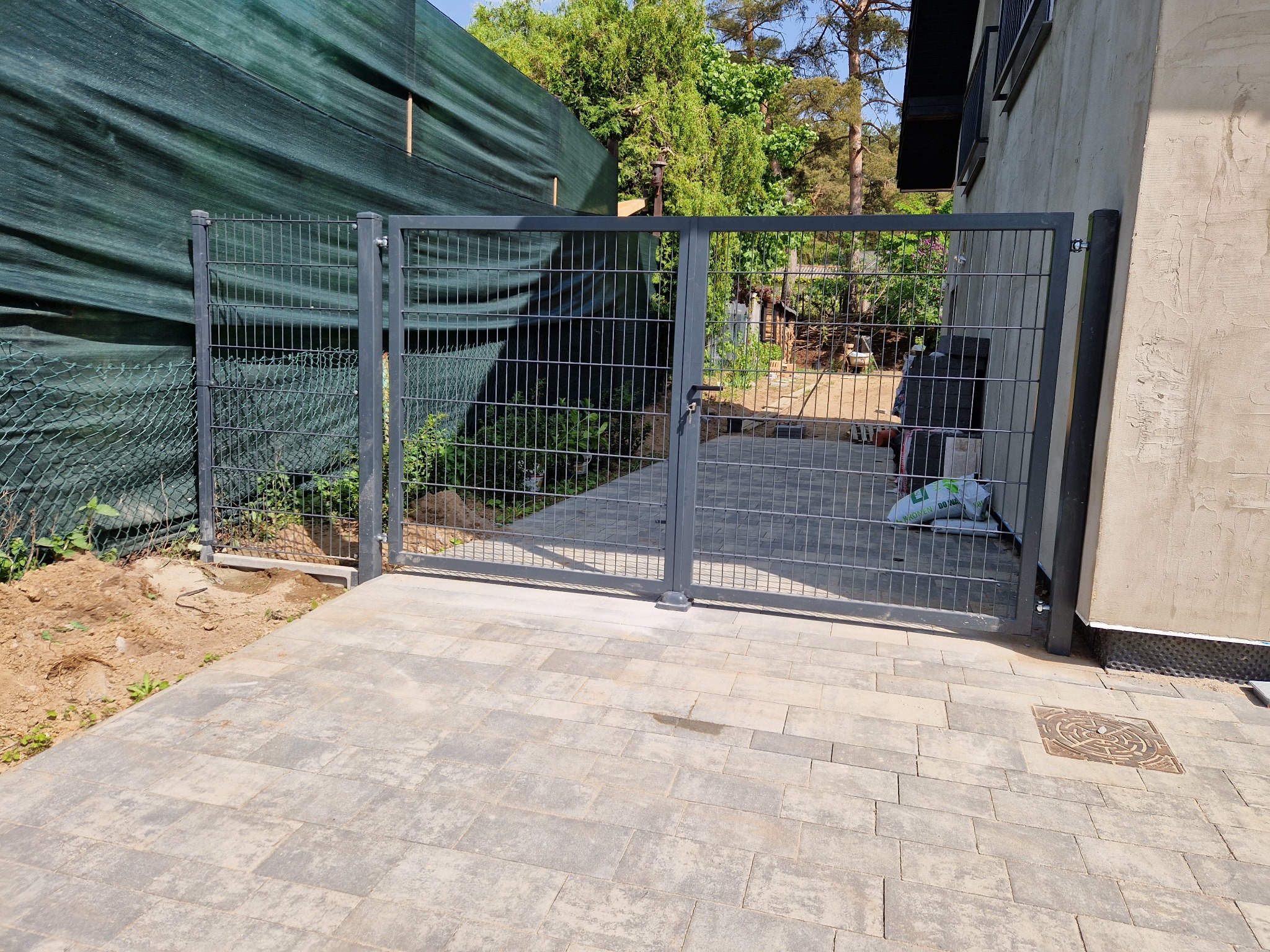 Szara, dwuskrzydłowa brama panelowa z niskim progiem, zamontowana na posesji z kostką brukową, widoczne worki budowlane w tle i zielona siatka maskująca po lewej stronie.