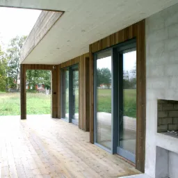 Zadaszony taras domu szkieletowego z drewnianą podłogą i ścianami, przesuwnymi szklanymi drzwiami oraz zewnętrznym kominkiem z cegły.