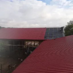 Częściowo pokryty czerwonym blachodachówką dach budynku gospodarczego, widoczny dekarz pracujący nad dalszym kryciem, odsłonięta więźba dachowa oraz folia paroizolacyjna.