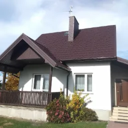 Dom jednorodzinny z brązowym dachem z dachówki, brązowymi elementami wykończenia, białymi ścianami i drewnianym tarasem z balustradą.