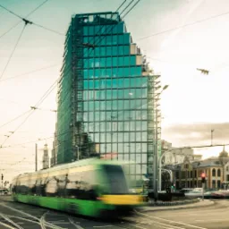 Szklany biurowiec w budowie z rusztowaniami, w tle tramwaj w ruchu i skrzyżowanie z sygnalizacją świetlną.