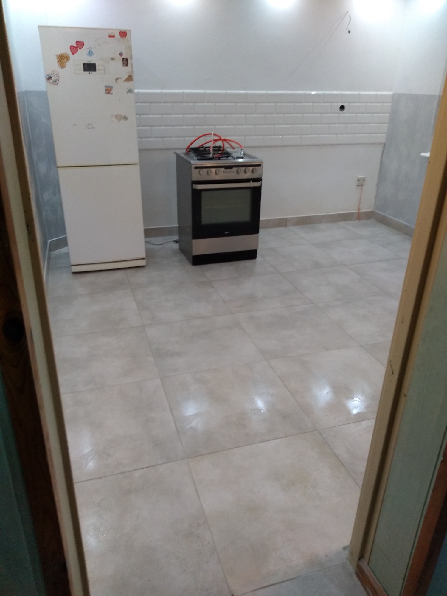 Wyremontowana kuchnia z białymi płytkami na ścianie, lodówką ozdobioną naklejkami i kuchenką gazową, widok zza framugi drzwi.