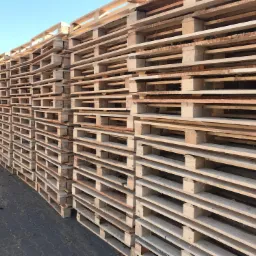 Stos jasnych, drewnianych palet ułożonych jeden na drugim, widoczne ślady użytkowania na niektórych paletach, szara kostka brukowa pod spodem, fragment ciemnego ogrodzenia w tle po lewej stronie...