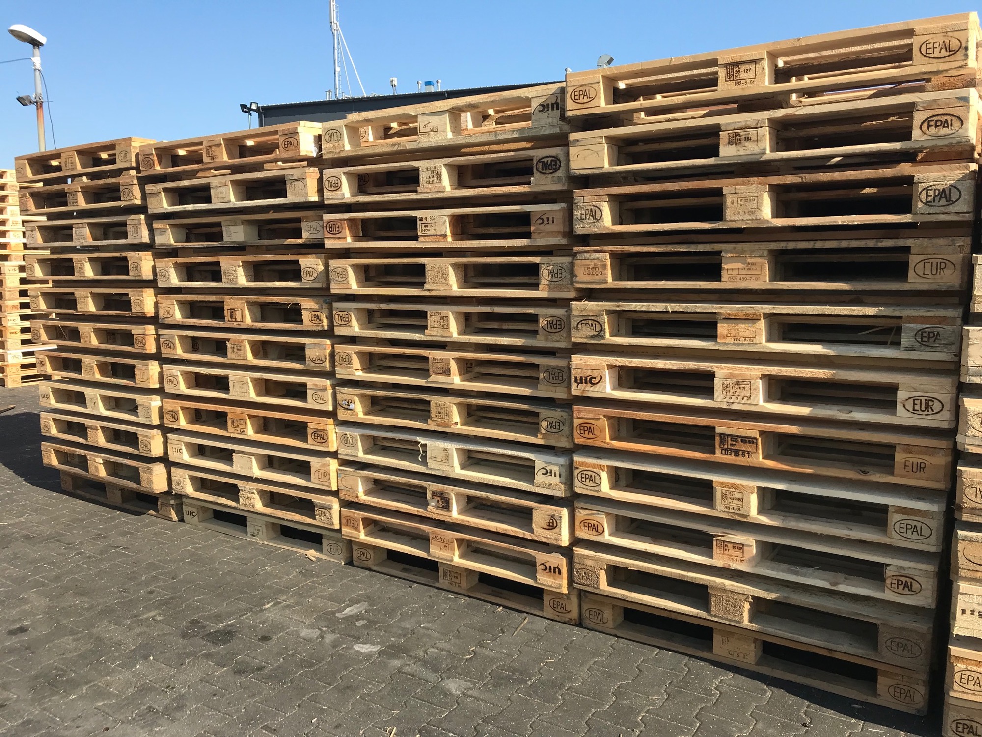 Stos jasnych, drewnianych palet EPAL ułożonych jedna na drugiej na szarym brukowanym podłożu, widoczne sygnatury i oznaczenia producentów na paletach, błękitne niebo w tle.
