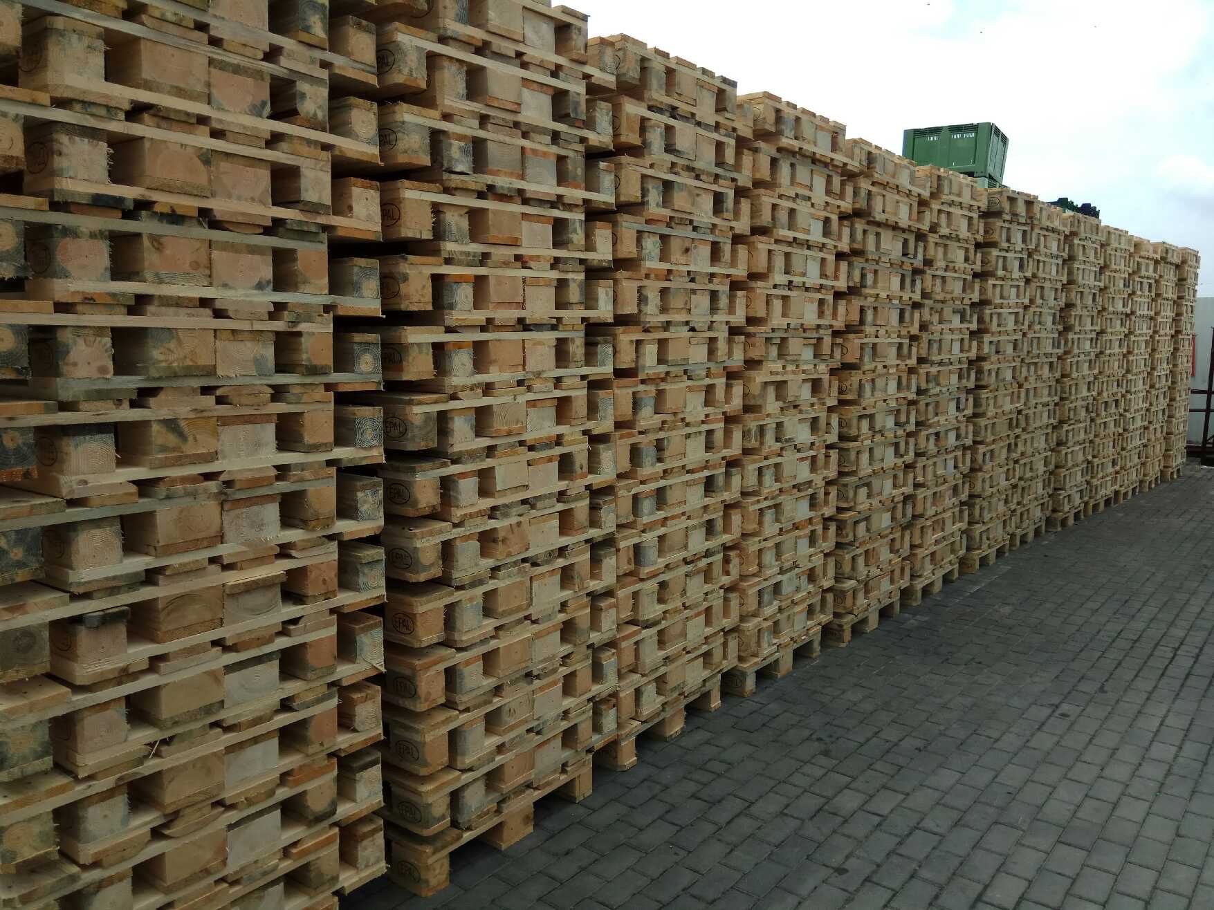 Stos drewnianych palet euro ułożonych w równy rząd na kostce brukowej, widoczny zielony pojemnik na górze stosu, pochmurne niebo w tle.