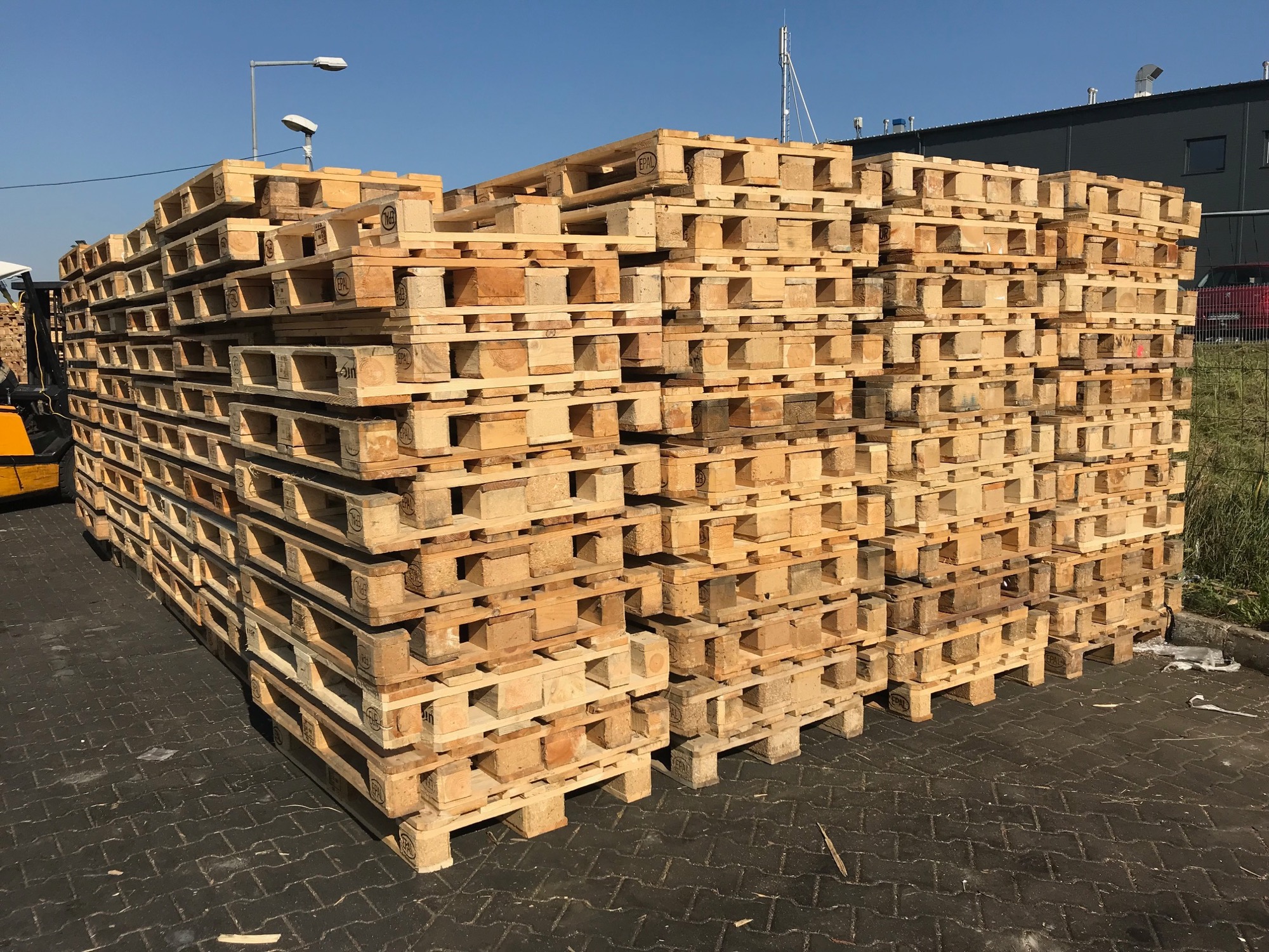 Stos drewnianych palet euro ułożonych na zewnątrz, widoczny wózek widłowy w tle, oświetlone przez słoneczne światło.