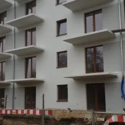 Nowe okna z brązowymi ramami w białym budynku wielorodzinnym z betonowymi daszkami nad oknami, widok z zewnątrz na plac budowy z rurami i barierkami.