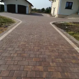 Ułożona, brązowo-żółta kostka brukowa tworząca podjazd do domu i garażu, obrzeża wykończone jaśniejszą kostką, widoczne budynki w tle, częściowo przygotowane rabaty.