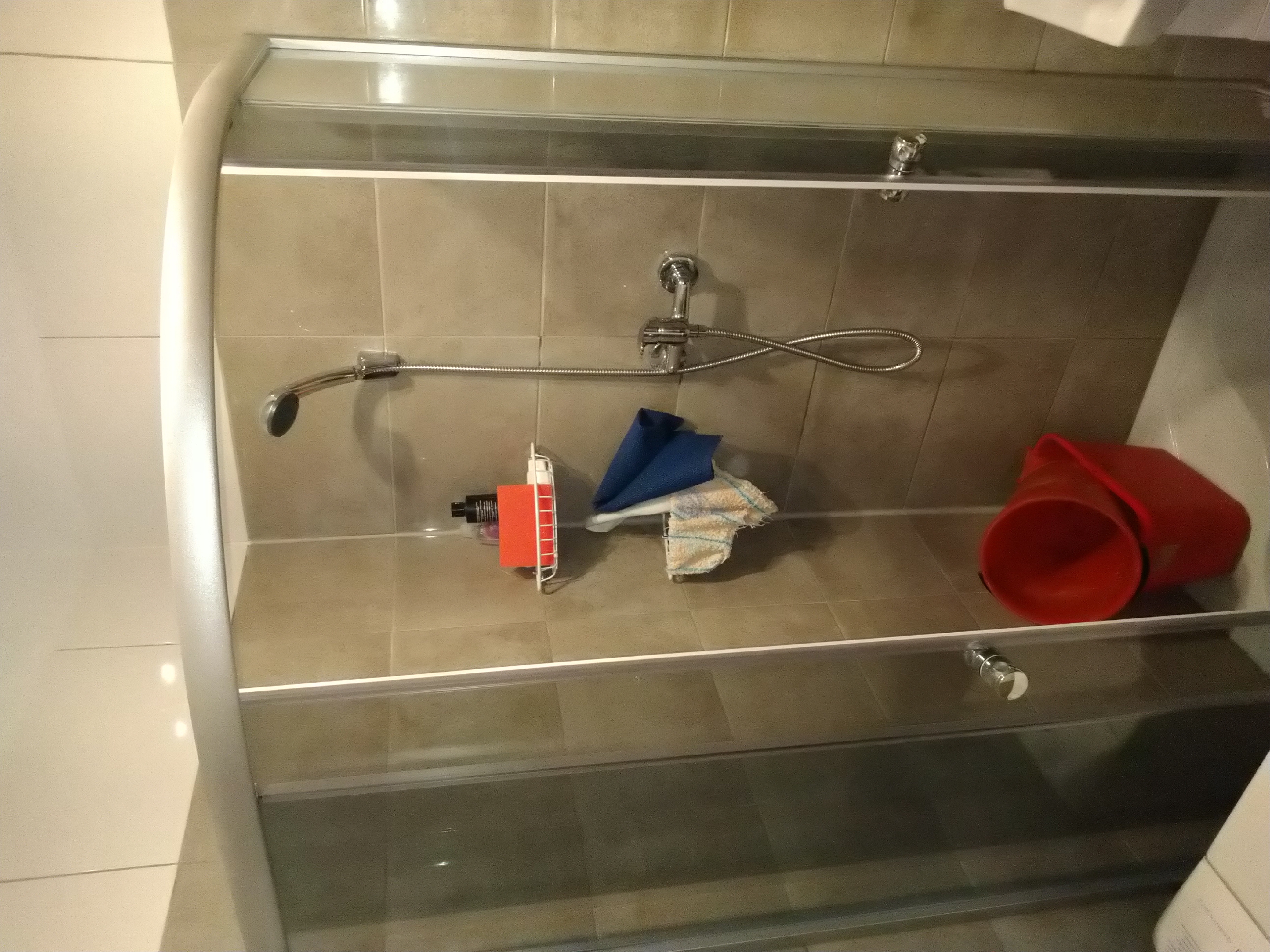 Wnętrze kabiny prysznicowej z beżowymi płytkami, widoczny zestaw prysznicowy, półka z organizerem, ściereczka, niebieska myjka oraz czerwone wiadro w rogu.