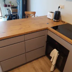 Fragment kuchni z drewnianym blatem, szarymi szafkami, piekarnikiem z wiszącą ścierką i płytą indukcyjną, w tle toster i czajnik elektryczny.