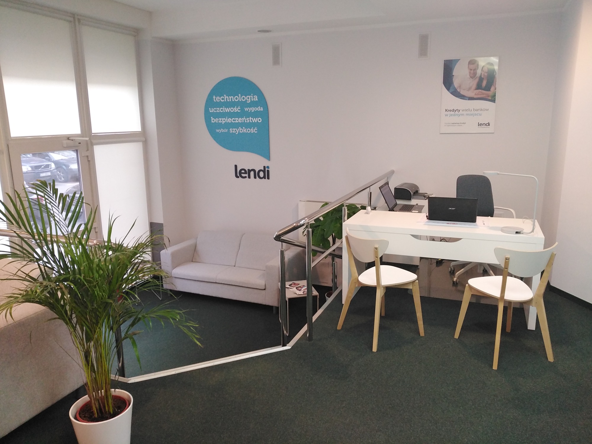 Jasne biuro z biurkiem, dwoma krzesłami, laptopem i rośliną doniczkową, logo firmy Lendi na ścianie i plakat reklamowy z hasłem 'Kredyty wielu banków w jednym miejscu'.
