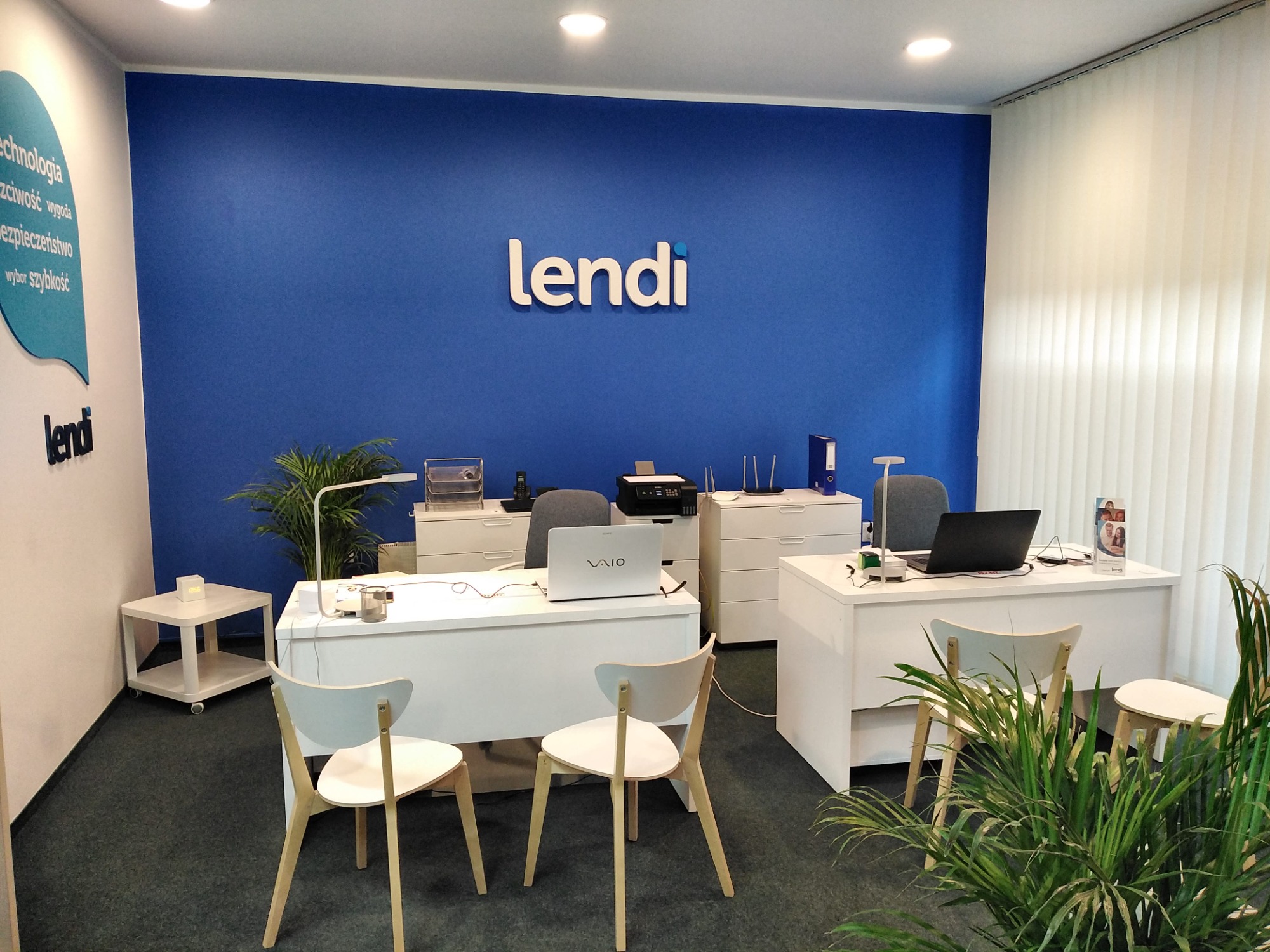 Nowoczesne biuro z logo 'lendi' na niebieskiej ścianie, białe biurka z laptopami i krzesłami, rośliny doniczkowe i rolety pionowe.