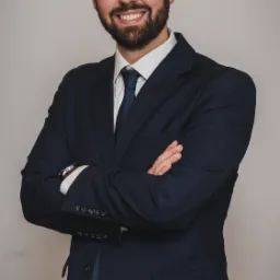 Uśmiechnięty mężczyzna z brodą w granatowym garniturze, białej koszuli i niebieskim krawacie, pozujący z założonymi rękami na tle jasnoszarej ściany, widoczny zegarek na nadgarstku.