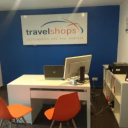 Wnętrze biura z białym biurkiem, komputerem, laptopem, pomarańczowymi krzesłami i logo 'travelshops - ogólnopolska sieć biur podróży' na niebieskiej ścianie.