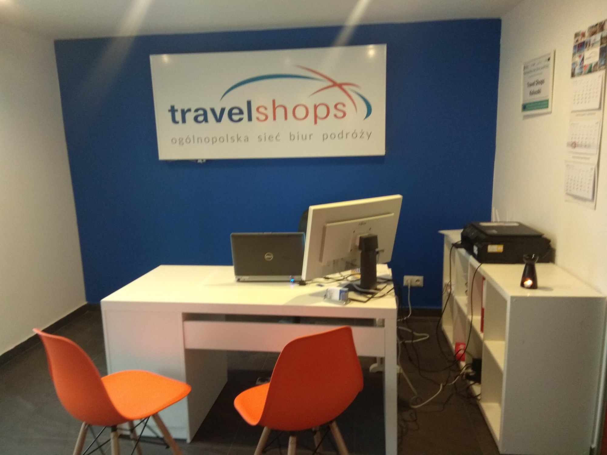 Wnętrze biura z białym biurkiem, komputerem, laptopem, pomarańczowymi krzesłami i logo 'travelshops - ogólnopolska sieć biur podróży' na niebieskiej ścianie.
