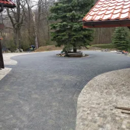 Szeroki podjazd wyłożony szarą kostką brukową, z okrągłym klombem wokół drzewa iglastego pośrodku, obrzeża z jasnej kostki, w tle budynek i drzewa.