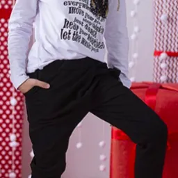 Dziewczynka w białej bluzce z długim rękawem i czarnych spodniach typu baggy, pozująca na tle świątecznych prezentów.