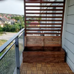 Drewniana zabudowa balkonu z ażurową ścianką i skrzynią na rośliny, widok na osiedle z balkonu wyłożonego panelami podłogowymi.