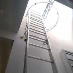 Metalowa drabina przymocowana do ściany, prowadząca do otworu w suficie z metalowym zabezpieczeniem, widok z dołu.