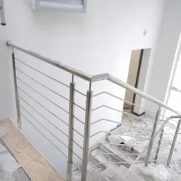 Nowoczesna balustrada ze stali nierdzewnej z poziomymi prętami, zamontowana na betonowych schodach w trakcie remontu, widoczne zabezpieczenia folią i okno z żaluzjami w tle.