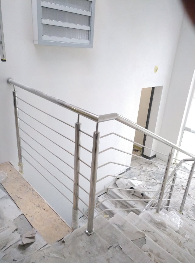 Nowoczesna balustrada ze stali nierdzewnej z poziomymi prętami, zamontowana na betonowych schodach w trakcie remontu, widoczne zabezpieczenia folią i okno z żaluzjami w tle.