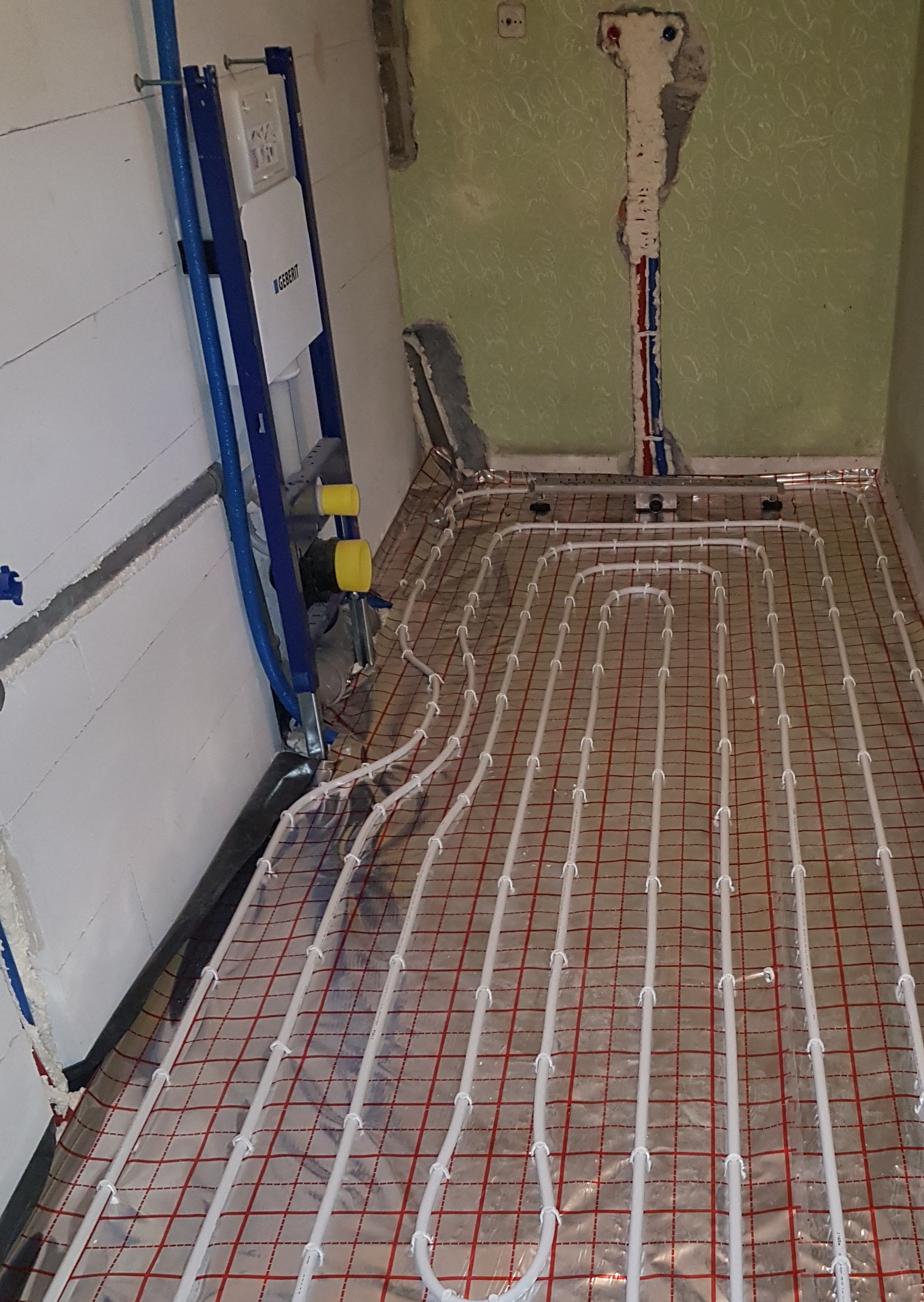 Instalacja ogrzewania podłogowego z rur PEX na folii aluminiowej z czerwoną siatką, widoczne elementy montażowe Geberit przy ścianie, surowy stan wykończenia wnętrza.