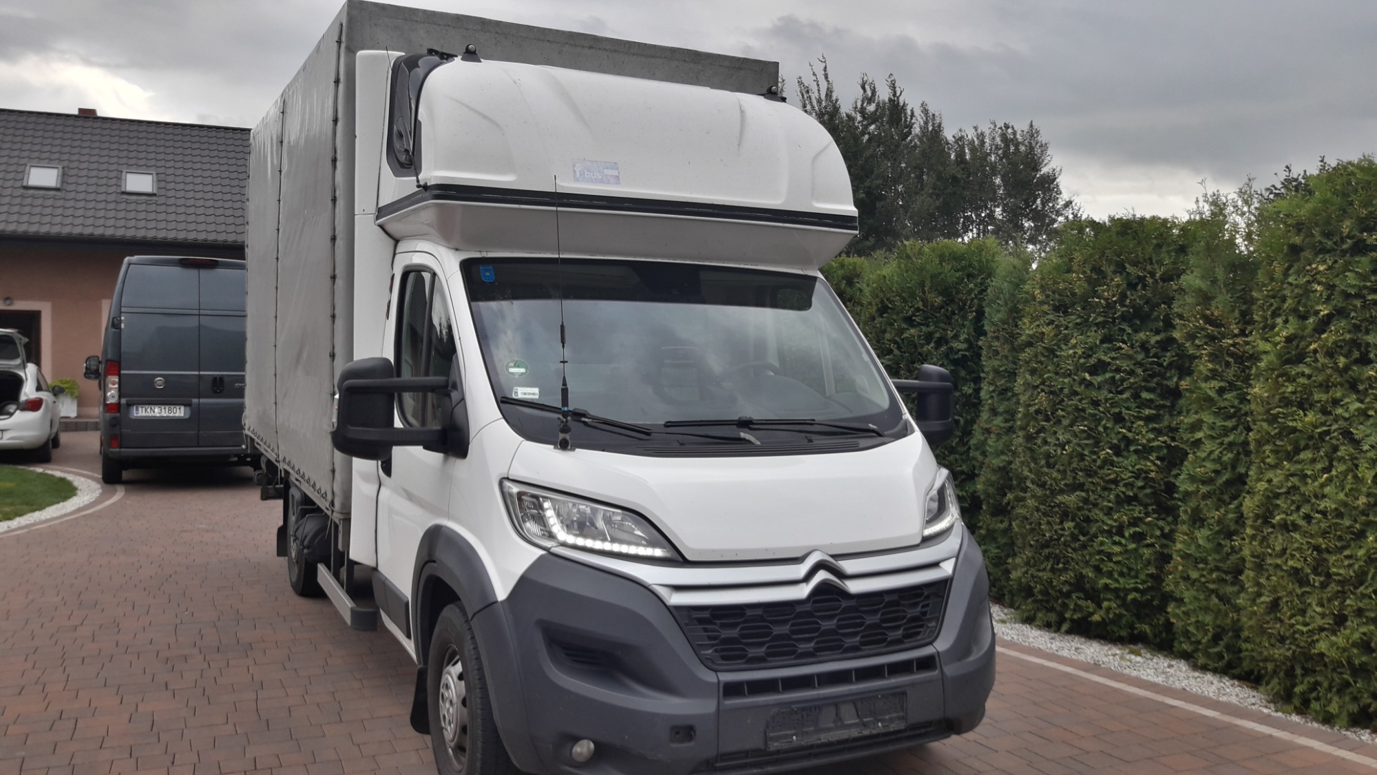 Biały samochód dostawczy z plandeką zaparkowany na brukowanej posesji, widoczny drugi bus w tle.