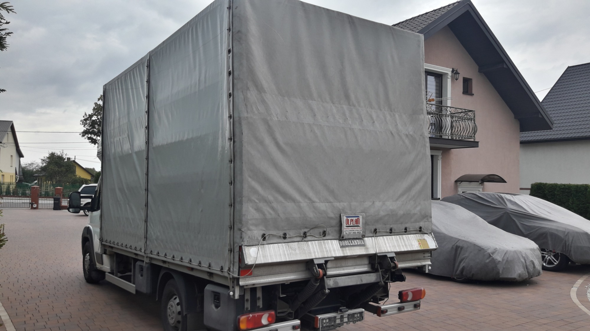 Tył samochodu dostawczego z plandeką na posesji z dwoma zaparkowanymi autami pod pokrowcami, widoczne logo firmy na plandece.