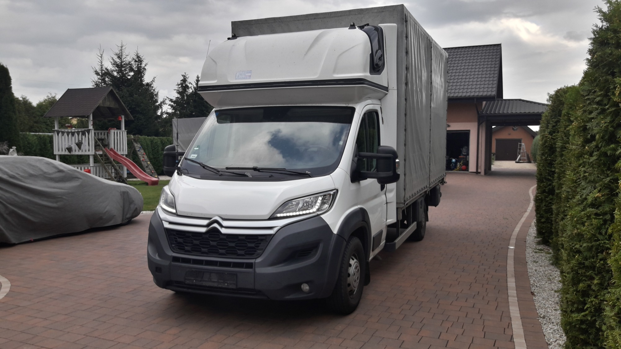 Biały bus z plandeką zaparkowany na posesji w Końskich, widoczny plac zabaw i dom w tle.