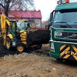 Florbud Ewelina Florjan - Żółta koparko-ładowarka Komatsu nabiera czarną ziemię, obok stoi zielona ciężarówka Volvo FM z żółto-czarnym pasem na zderzaku, w tle ceglane budynki z czerwonymi dachami.
