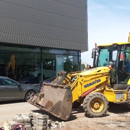 Florbud Ewelina Florjan - Żółta koparko-ładowarka Komatsu stoi na placu budowy przed nowoczesnym budynkiem biurowym z dużymi oknami, obok leży stos betonowej kostki brukowej, a w tle widoczny jest szary samochód osobowy.