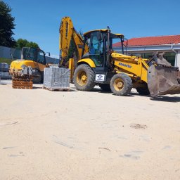 Florbud Ewelina Florjan - Żółta koparko-ładowarka Komatsu na placu budowy obok palet z kostką brukową i mniejszej koparki, widoczny budynek z czerwoną dachówką.