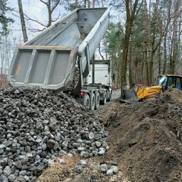 Florbud Ewelina Florjan - Wywrotka z podniesioną skrzynią wysypuje kamienie na ziemię, obok pracuje żółta koparka Komatsu, w tle las.