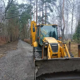 Żółta koparko-ładowarka Komatsu na żwirowej drodze w lesie, widoczne świeżo wysypane kamienie.