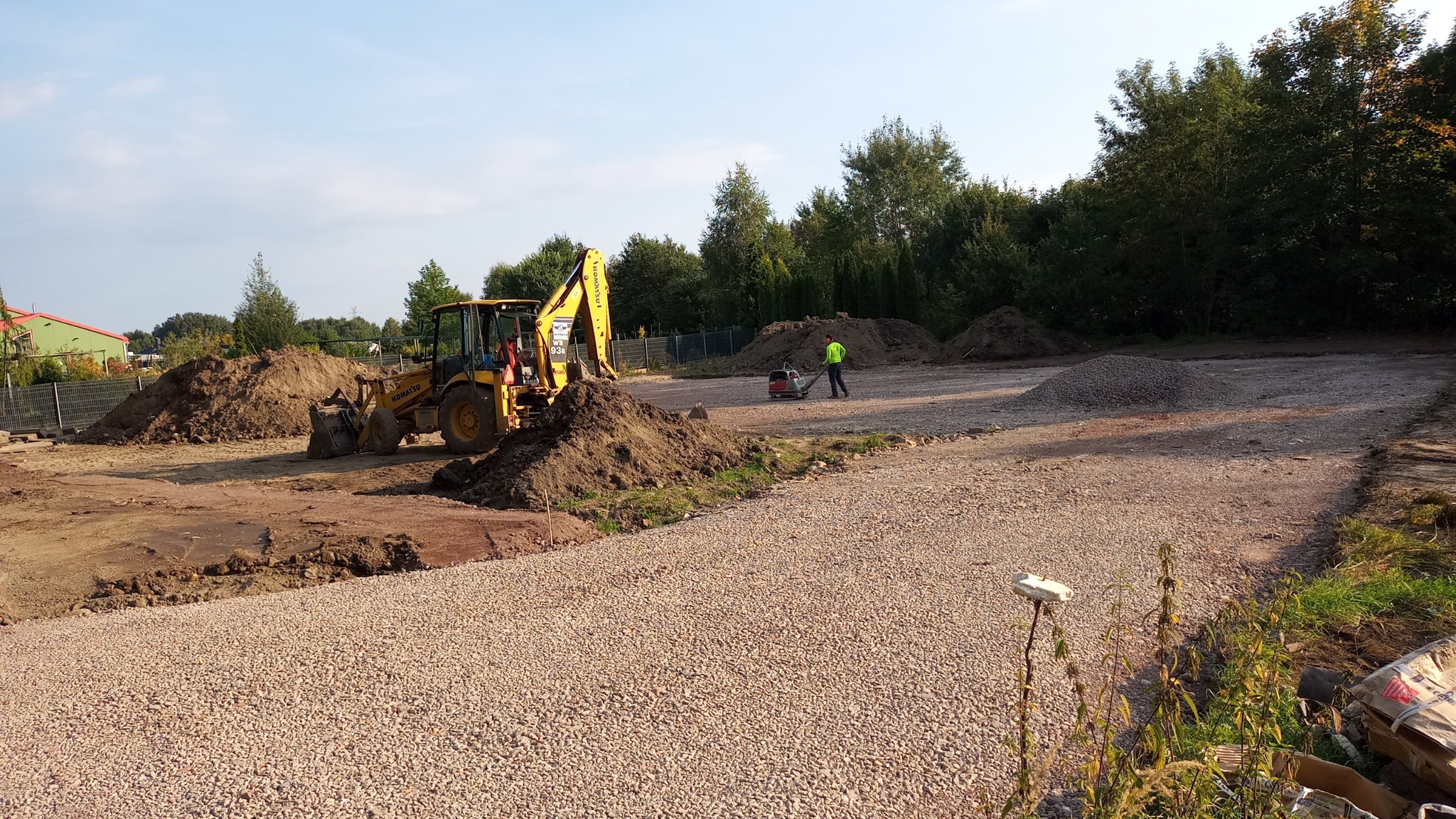 Żółta koparko-ładowarka Komatsu obok pryzmy ziemi na placu budowy, w tle pracownik z zagęszczarką i hałdy żwiru.