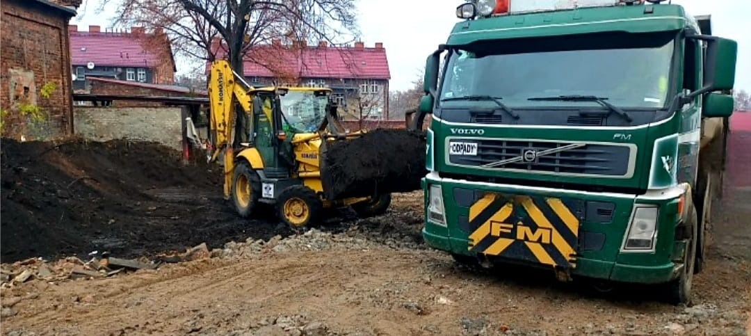 Żółta koparko-ładowarka Komatsu nabiera czarną ziemię, obok stoi zielona ciężarówka Volvo FM z żółto-czarnym pasem na zderzaku, w tle ceglane budynki z czerwonymi dachami.