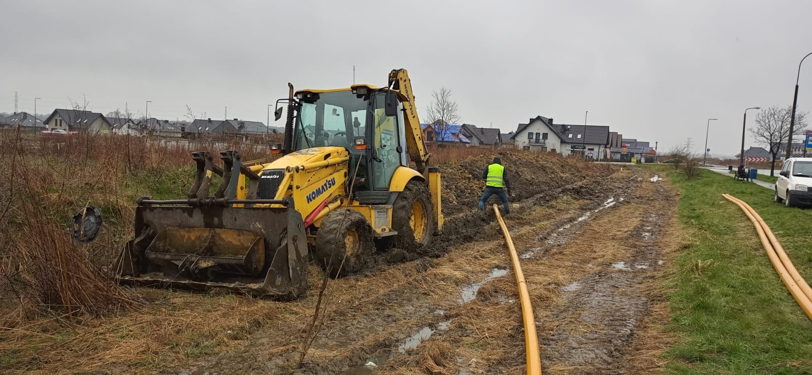 Żółta koparko-ładowarka Komatsu w błotnistym terenie z pracownikiem w kamizelce odblaskowej układającym pomarańczowe rury, w tle zabudowa jednorodzinna i pochmurne niebo.
