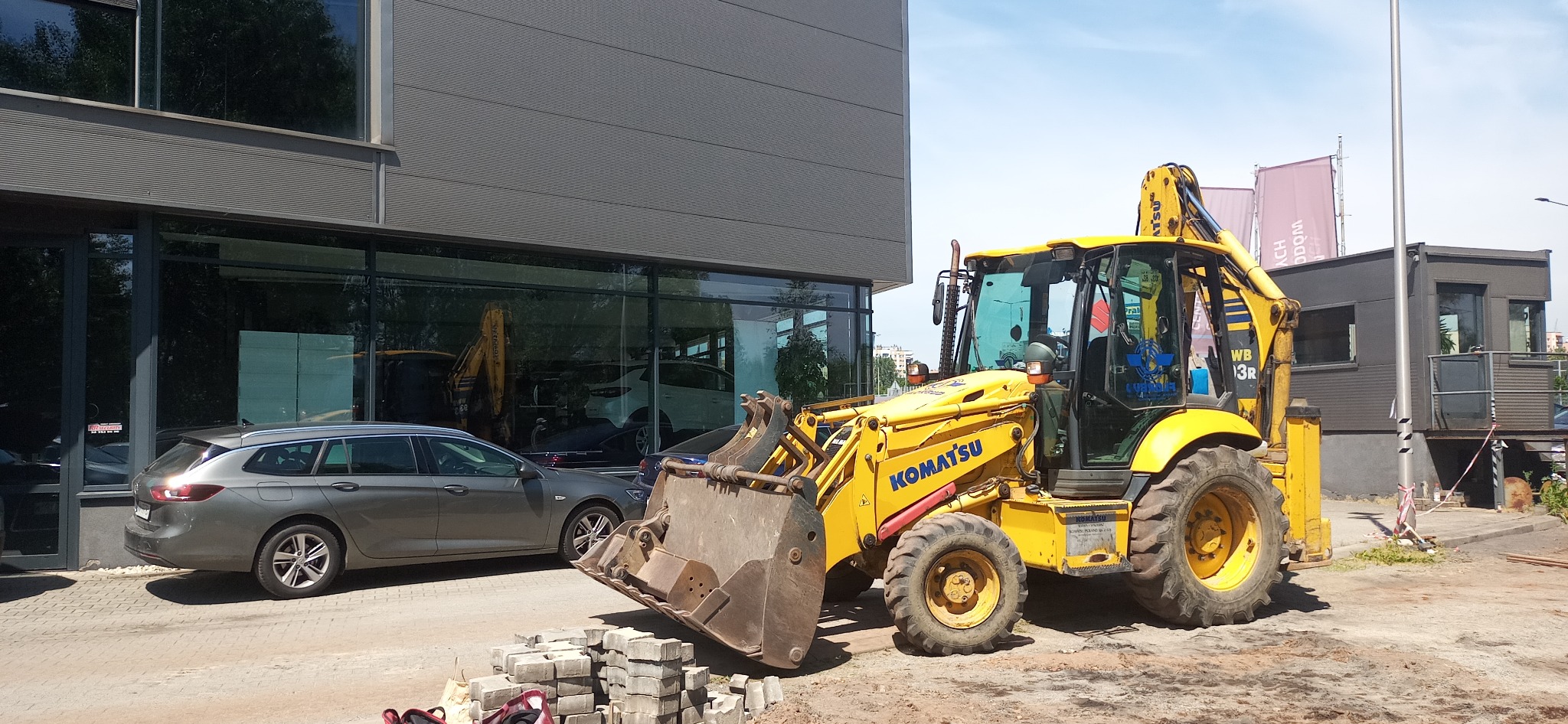 Żółta koparko-ładowarka Komatsu stoi na placu budowy przed nowoczesnym budynkiem biurowym z dużymi oknami, obok leży stos betonowej kostki brukowej, a w tle widoczny jest szary samochód osobowy.