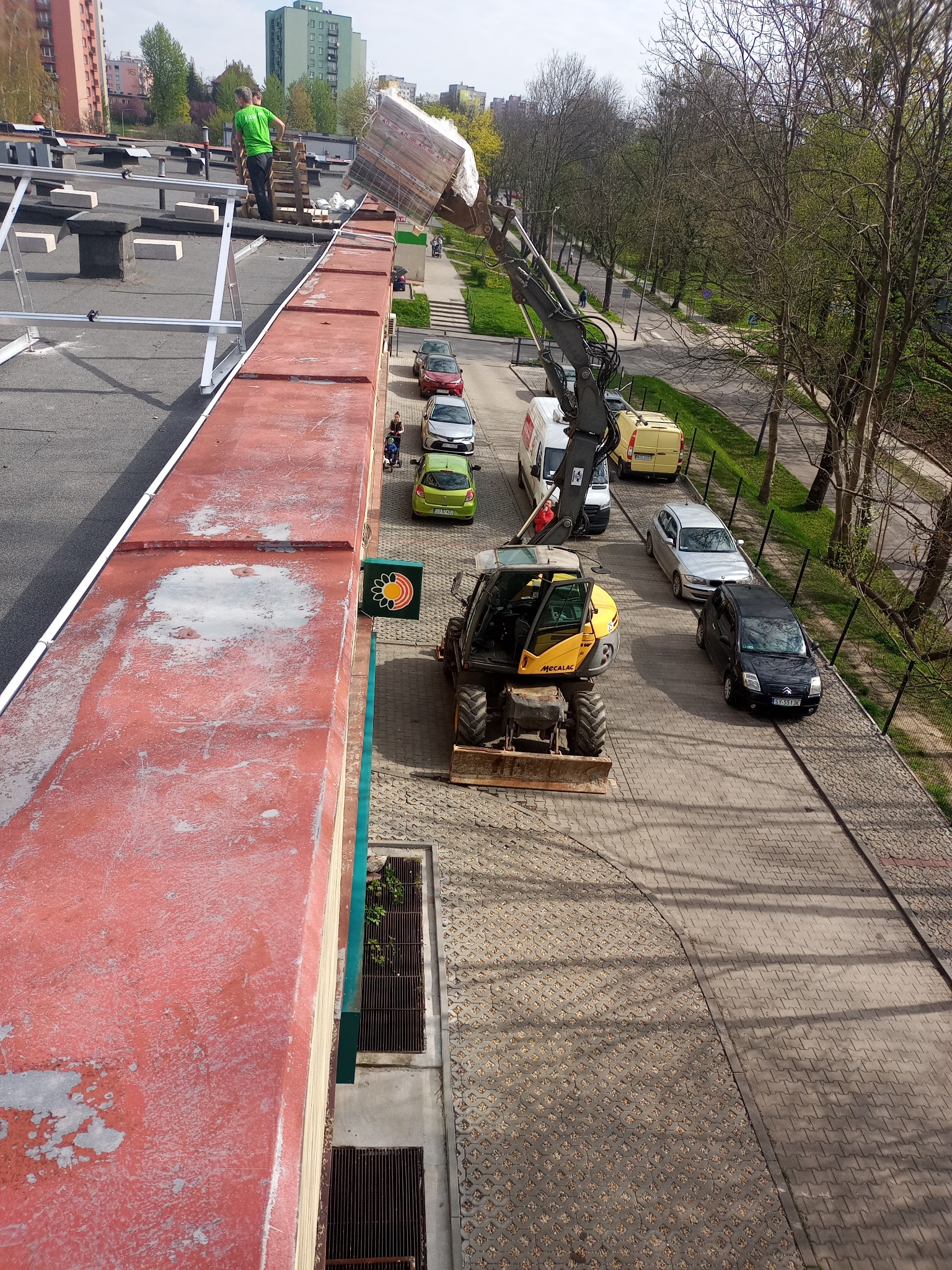 Żółta koparko-ładowarka z wysięgnikiem podnosi pakiet materiałów na dach budynku, gdzie stoi pracownik w zielonej koszulce, widok z góry na chodnik, samochody i drzewa w tle.