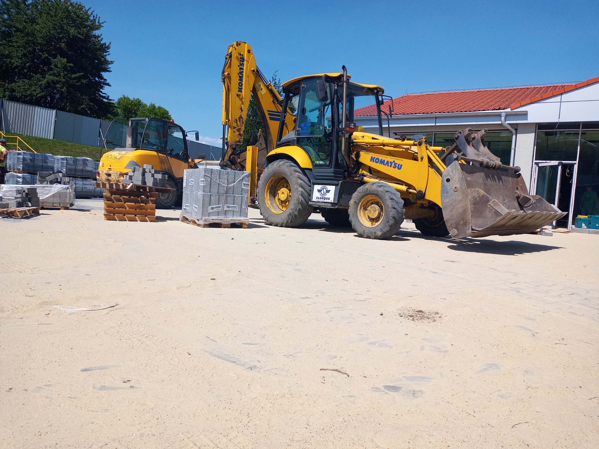 Żółta koparko-ładowarka Komatsu na placu budowy obok palet z kostką brukową i mniejszej koparki, widoczny budynek z czerwoną dachówką.
