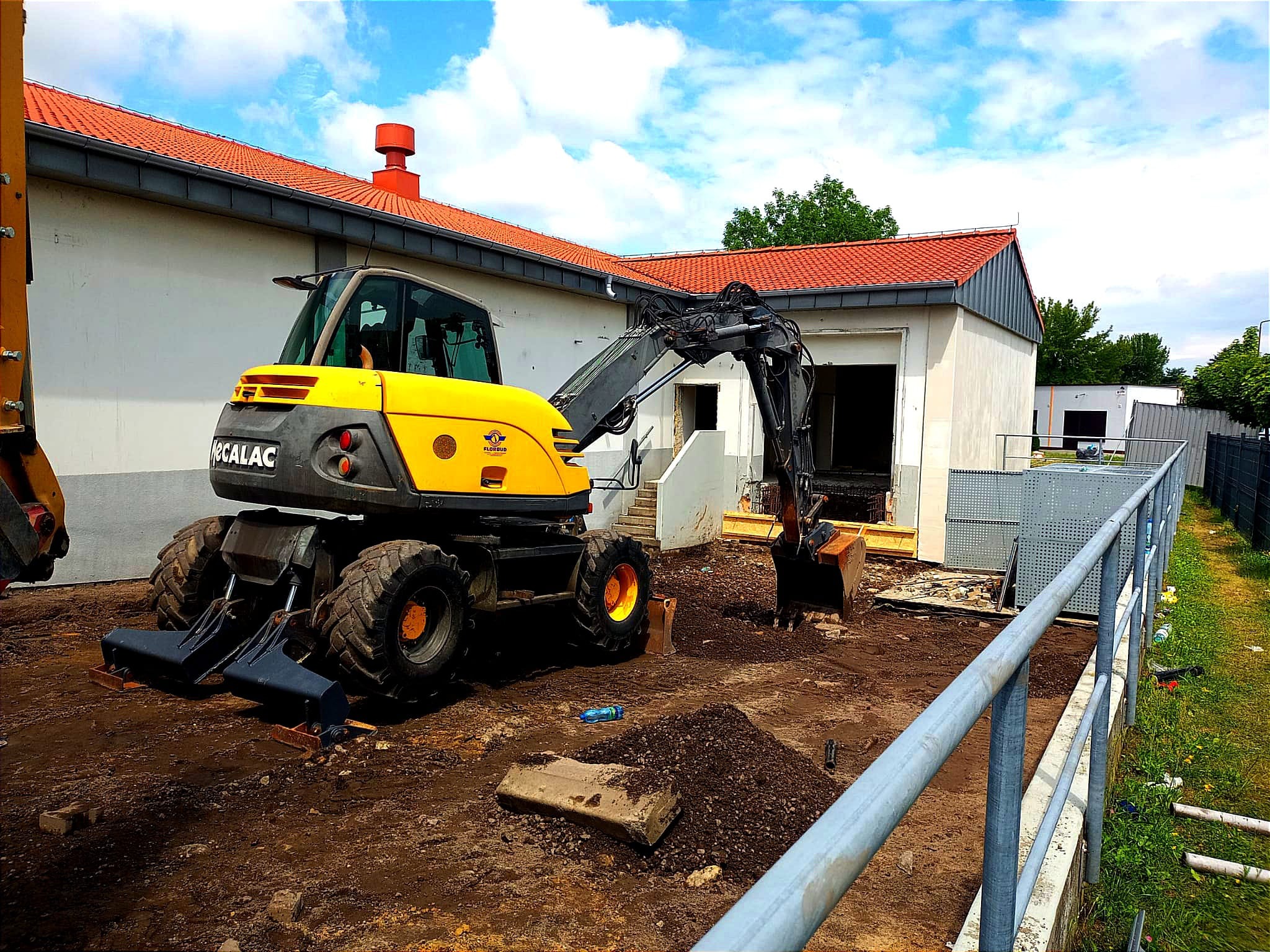Żółto-czarna koparka kołowa MOCALAC na placu budowy, z wykopem przy budynku z czerwonym dachem, w tle błękitne niebo z chmurami.