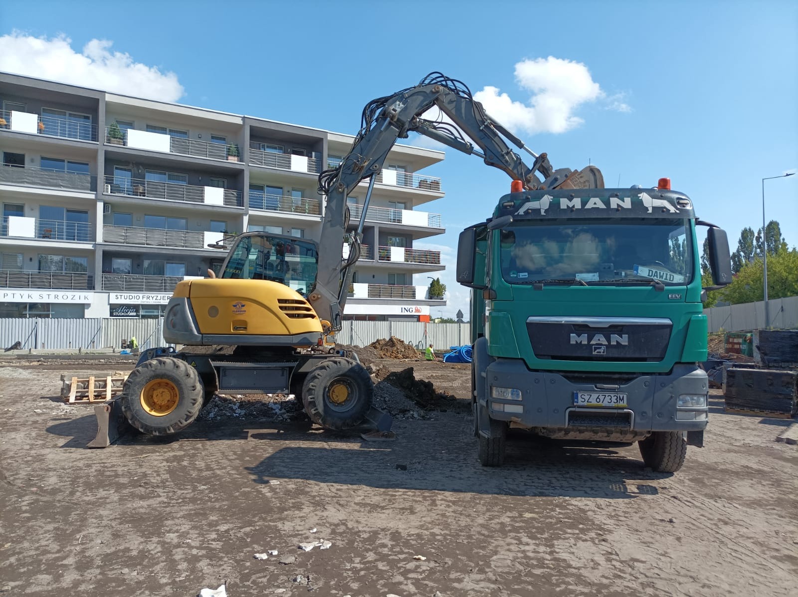 Żółta koparka kołowa ładuje urobek na zieloną ciężarówkę MAN na placu budowy, w tle budynek mieszkalny i błękitne niebo.