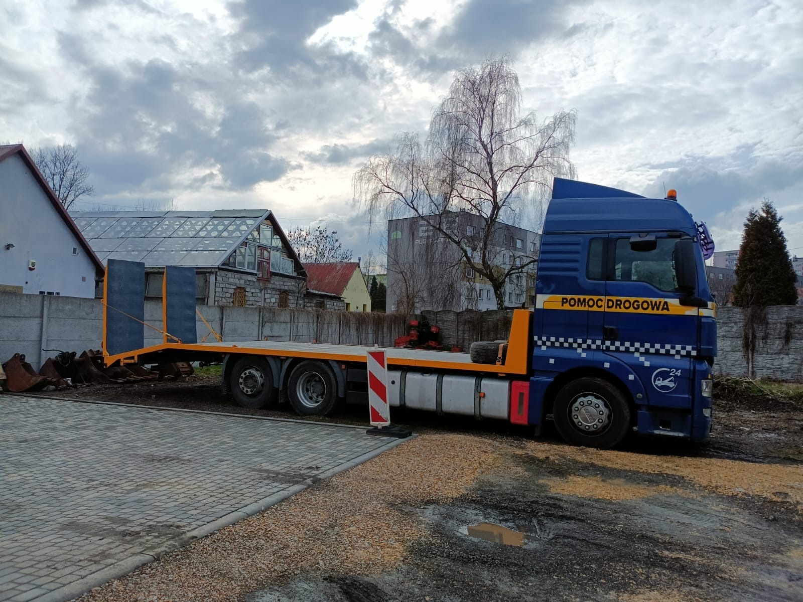 Niebieska laweta z napisem 'Pomoc Drogowa' zaparkowana na brukowanym placu, z łyżkami do koparki na platformie, tło stanowi zabudowa miejska pod pochmurnym niebem.
