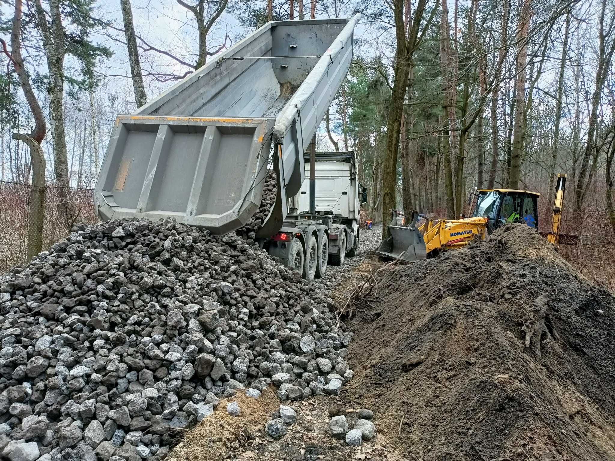 Wywrotka z podniesioną skrzynią wysypuje kamienie na ziemię, obok pracuje żółta koparka Komatsu, w tle las.