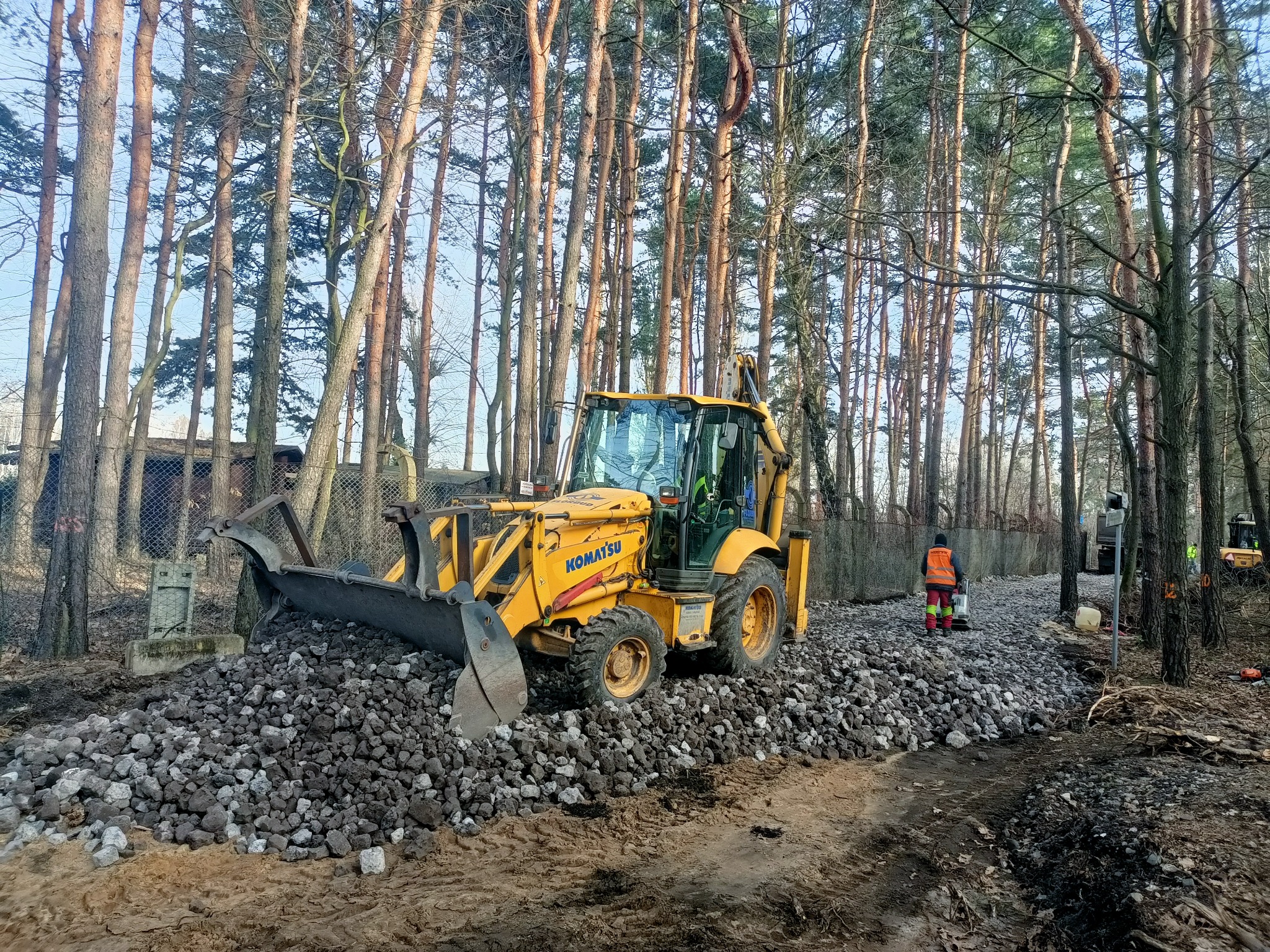 Żółta koparko-ładowarka Komatsu na usypanym żwirowym podłożu w lesie, pracownik w pomarańczowej kamizelce zagęszcza podłoże ubijarką, w tle wysokie drzewa i siatka ogrodzeniowa.